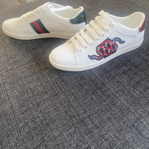 Gucci Ace skor - Gucci Ace sneakers i vitt skinn med klassiska gröna och röda ränder på ena sidan och broderad röd orm på andra. Baksidan har metallicröd och metallicgrön detalj med ormskinnsmönster och Gucci-logga. Snygga snörade sneakers med rund tå och platt sula.