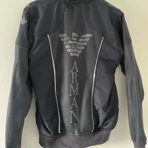 Svart bomberjacka från Armani, M - Svart bomberjacka från Armani med stora logotyper och örntryck på ryggen. Jackan har mesh-detaljer på ärmar och sidor, dragkedja framtill och ribbade muddar. Snygg sportig look med vita kontrastlinjer och hög krage. Perfekt för dig som gillar streetwear och vill sticka ut.