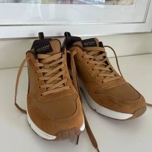 Bruna sneakers från Skechers - Snygga bruna sneakers från Skechers med ovandel i mocka och syntet, vita detaljer på sulan och bruna snören. Skorna har memory foam-innersula för extra komfort och en sportig, chunky sula. Perfekta för dig som gillar streetstyle och vill ha bekväma skor hela dagen. Helt nya!! Nypris: 1499kr