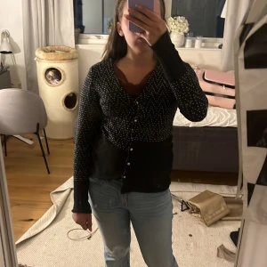 Svart kofta med vita prickar och knappar - Svart kofta med vita prickar i zigzag-mönster och snygga pärlemorknappar framtill. Långärmad modell med ribbade muddar och figurnära passform. Perfekt att slänga över en topp för en chill men ändå stilsäker look.