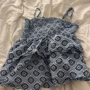 Blå mönstrad linnetopp Gina Tricot - Supersöt blå linnetopp från Gina Tricot med smala axelband och volangdetalj. Toppen har ett unikt mönster i marinblått och vitt, smockad överdel och luftig passform. Perfekt för varma dagar och ger en fräsch sommarkänsla.