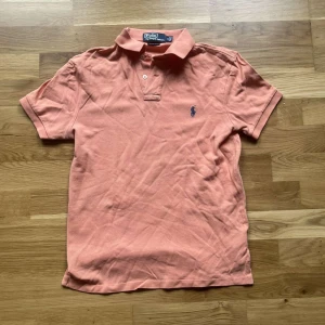Ralph Lauren piké  - Snygg korallfärgad pikétröja från Ralph Lauren med klassisk krage, korta ärmar och blå broderad logga på bröstet. Tröjan är i bomull och har knappar vid halsen. Perfekt för en chill och stilren look. Fint skick förutom att färgen är lite blekt 
