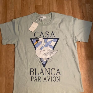 Ljusblå Casablanca t-shirt med svan - Casablanca t-shirt i ljusblått med stor grafisk print framtill av en svan och texten 'Casa Blanca Par Avion'. Klassisk passform, rund hals och korta ärmar. Tillverkad i mjuk bomull, perfekt för en chill och stilren look.