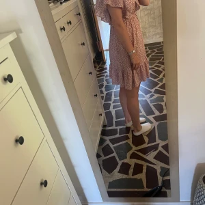 Rosa blommig omlottklänning - Supersöt kort rosa klänning med vitt blommönster och volangkant. Omlottmodell med v-ringning och rosett knyt vi midjan som ger en snygg look. Lätta, korta ärmar och flowigt tyg – perfekt för varma dagar. Köptes i butik i Göteborg men kommer inte till användning vilket är synd då den är så fin!💖