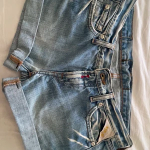 Ljusblå jeansshorts från replay - Snygga ljusblå jeansshorts från AOH Denim med slitna detaljer och uppvikta benslut. Shortsen har klassiska fickor fram och bak, coola kontrastsömmar och en läderpatch med logga bak. Perfekta för varma dagar och en avslappnad stil.