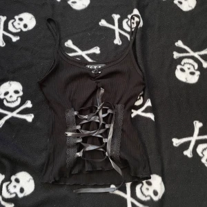 Svart handsydd korsettlinne med snörning  - Supersnygg svart korsettinspirerad linne-topp från Stay i ribbad bomull. Framsidan har bred snörning med satinband och spetsdetaljer längs kanterna. Smala axelband och croppad passform ger en edgy vibe. Perfekt för dig som gillar alternativ stil. Plagget är handsytt, vilket innebär att det kan förekomma mindre skavanker tex lösa trådar! 🪡 