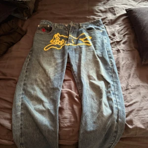 Icecream jeans med gul grafik - Unika blå jeans från Icecream med fet gul grafik framtill och broderade bokstäver på bakfickorna. Klassisk femficksmodell i denim med raka ben och normal passform. Perfekta för dig som vill sticka ut med streetwear-vibe.