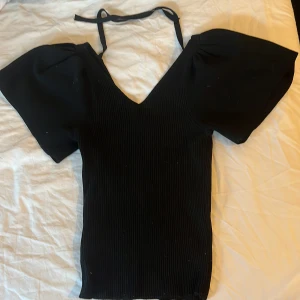 Svart ribbad topp med puffärm från ONLY - Svart ribbad topp från ONLY med v-ringning, korta puffärmar och knytband i nacken. Snygg croppad passform som ger en trendig siluett. Perfekt för dig som gillar stilrena och coola detaljer. Snören där bak för att knyta en rosett. 220kr plus frakt