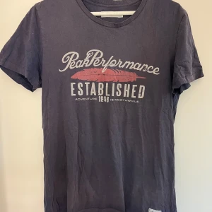 Mörkblå Peak Performance t-shirt S - Snygg mörkblå t-shirt från Peak Performance med tryck framtill i vitt och rött. Klassisk rund hals och korta ärmar. Skön passform och tillverkad i mjuk bomull, perfekt till jeans eller shorts.