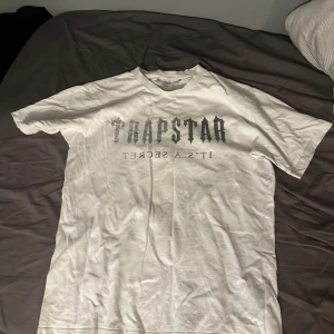 Vit Trapstar t-shirt med tryck - Vit t-shirt från Trapstar med svart tryck på bröstet där det står 'TRAPSTAR IT'S A SECRET' i gotisk stil. Klassisk passform med rund hals och korta ärmar. Tillverkad i mjuk bomull, perfekt för en clean streetwear-look. Passar m och L