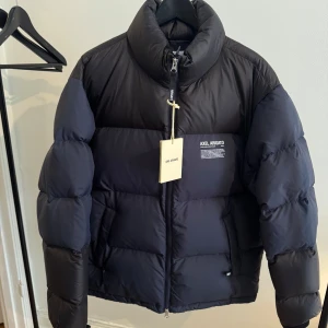 Svart Blå dunjacka från Axel Arigato - Snygg svartblå dunjacka från Axel Arigato. Jackan har dragkedja framtill och är riktigt fluffig och varm, perfekt för kalla dagar. Stilren design som ger en cool vibe till din outfit. Nypris: 3000kr