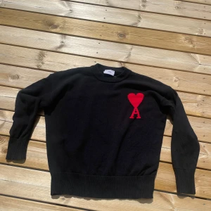 Svart stickad tröja från Ami Paris - Svart stickad tröja från Ami Paris med röd hjärtlogga och bokstaven A broderad på bröstet. Klassisk rund halsringning, ribbade muddar och nederkant. Perfekt för dig som gillar stilrena plagg med en twist. Skickar inom 24h.
