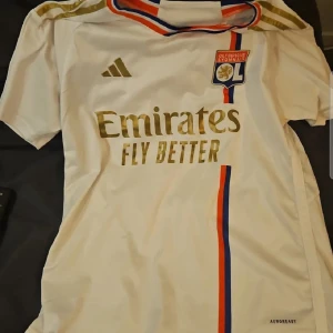Vit Olympique Lyon Adidas matchtröja,lite skador som man kan se på bilden - Snygg vit fotbollströja från Olympique Lyonnais med guldiga Adidas-detaljer och Emirates-tryck. Tröjan har korta ärmar, röd och blå rand längs framsidan och klubbmärke på bröstet. Tillverkad i ventilerande polyestermaterial (AEROREADY).