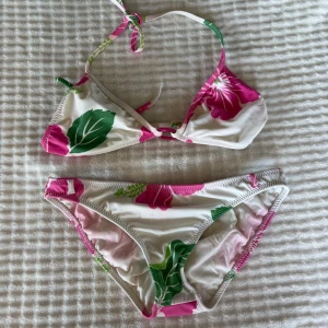Blommig bikini från Raisins, M - Passar S-M passar mig som har storlek S ungefär! Snygg bikini från Raisins i storlek M med blommigt mönster i rosa, grönt och vitt. Triangeltopp med knyt i nacken och klassiska bikinitrosor. Perfekt för stranden eller poolen, med tropiska vibbar och sköna färger.