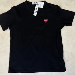 Svart t-shirt Comme des Garçons Play - Svart t-shirt från Comme des Garçons Play med klassiskt rött hjärta med ögon broderat på bröstet. Enkel och stilren design, rund hals och korta ärmar. Tillverkad i mjuk bomull som känns skön mot huden. Perfekt för dig som gillar streetstyle och ikoniska detaljer. Det är rrreepp