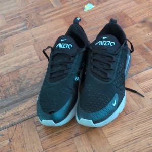 Nike Air Max 270 svarta sneakers - Svarta Nike Air Max 270 sneakers med vit sula och tydlig Air Max-bubbla i hälen. Ovandelen är i mesh och syntet, med snörning och diskreta vita loggor. Skorna har en sportig vibe och passar perfekt till streetwear eller träning.