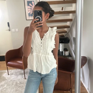 Vit blus med volang och spetsdetaljer - Supersöt vit blus med volang och spetsdetaljer längs axlarna. Blusen är ärmlös, har knappar framtill och ett luftigt, flowy tyg som ger en romantisk vibe. Perfekt för varma dagar eller när du vill känna dig extra fin.