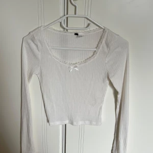 Vit ribbad långärmad topp med spets - Supersöt vit ribbad långärmad topp från H&M med spetskant runt urringningen och en liten rosett framtill. Croppad modell som sitter snyggt och har en feminin vibe. Perfekt att styla med jeans eller kjol för en trendig look.