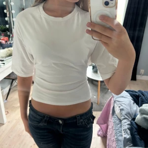 Vit croppad topp med knyt från Nelly - Säljer en vit croppad topp från Nelly med korta ärmar och rund hals. Toppen har öppen rygg och snygg knytning bak som ger en cool och trendig look. Perfekt till jeans eller kjol för en fräsch stil.