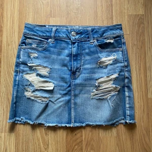 Blå jeanskjol från American Eagle - Snygg blå jeanskjol från American Eagle med slitna detaljer och fransig nederkant. Klassisk femficksmodell med dragkedja och knapp. Gjord i stretchigt denim för skön passform. Är i stolek us 10 vilket motsvarar ca 43 i europeiska storlekar men jag personligen skulle säga att den passar som en S/M. Hör gärna av er vid frågor!💕