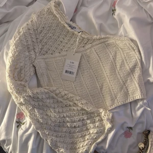 Offwhite stickad oneshoulder topp NA-KD - Superfin offwhite stickad topp från NA-KD med oneshoulder-design och lång ärm. Toppen har ett luftigt, virkat mönster som ger en cool och trendig vibe. Perfekt att styla med höga jeans eller kjol för en snygg look.