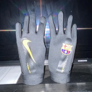 Nike x FC Barcelona svarta handskar - Svarta Nike Therma-Fit handskar med gult swoosh och FC Barcelona-logga. Slimmad passform med grepp på insidan av handflatan och touchscreen-kompatibla fingertoppar. Perfekta för fotbollsträning eller kalla dagar på läktaren.