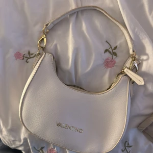 Vit axelväska från Valentino - Snygg vit axelväska från Valentino med guldfärgade detaljer och dragkedja. Väskan har en stilren, rundad form och är gjord i strukturerat skinnliknande material. Perfekt storlek för det viktigaste och ger en clean look till din outfit.