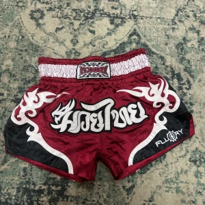 Vinröda Muay Thai shorts från Fluory - Snygga Muay Thai shorts från Fluory i vinrött med vita och svarta detaljer samt coolt mönster och broderad text framtill. De har bred resår i midjan och är tillverkade i glansigt syntetmaterial som ger maximal rörelsefrihet under träning eller match.