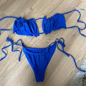 Kornblå bikini med knytband, stl L - Snygg blå bikini med knytband både upptill och nedtill. Går att knyta överdelen på olika sätt. scrunch-detaljer. Tillverkad i stretchigt material med 80% nylon och 20% spandex. Perfekt för stranden eller poolen i sommar!