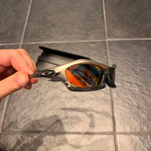 Oakley Splice solglasögon silver/orange - Oakley Splice solglasögon med futuristisk design, silverfärgad båge och spegelblanka orange linser. Snyggt sportig look med markerade detaljer och Oakley-logga på skalmarna. Perfekta för dig som vill sticka ut med en unik stil.