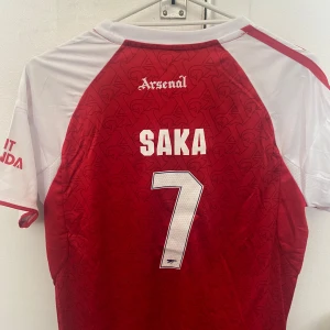 Arsenal Saka 7 matchtröja Adidas - Riktigt snygg Arsenal fotbollströja med Saka 7 på ryggen. Tröjan är röd med vita ärmar och detaljer, Adidas-logga och klubbmärke på bröstet samt Champions League-märke på ärmen. Tillverkad i lätt och ventilerande material, perfekt för match eller träning.