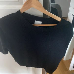 Svart croppad stickad topp H&M - Svart croppad stickad topp från H&M med korta ärmar och rund halsringning. Enkel och stilren design som är lätt att matcha. Perfekt för dig som gillar en clean look med lite edge.
