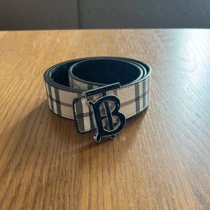 Rutig Burberry bälte med B-spänne - Snyggt bälte från Burberry med klassiskt rutigt mönster i beige, svart och brunt. Bältet har ett stort metallspänne med Burberrys ikoniska B-logga. Perfekt accessoar för att lyfta din outfit med en lyxig touch.