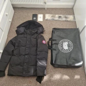 Canada Goose jacka - Säljer en svart dunjacka från Canada Goose i storlek S. Jackan har huva, klassisk logga på ärmen och stängs med både dragkedja och knappar. Fodrad med dun och fjädrar för maxad värme. Perfekt för kalla dagar och riktigt snygg streetstil.