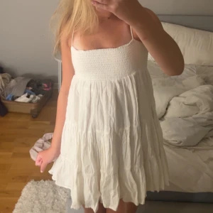 Vit somrig kortklänning från Zara - Supersöt vit kortklänning från Zara med smock upptill och tunna axelband. Klänningen har en luftig, volangig kjol och är perfekt för varma dagar. Tillverkad i bomull för en skön och lätt känsla.