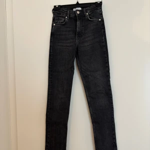 Svarta jeans med slits från Zara - Snygga svarta jeans med slits från Zara med hög midja och råa, slitsade benslut. Jeansen har klassisk femficksdesign, dragkedja och knapp framtill. Perfekta för dig som gillar en tight passform och coola detaljer. Finns ett hål (se sista bilden).