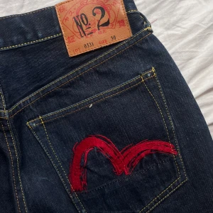 Evisu jeans med röd brodyr, stl 30 - Säljer ett par klassiska Evisu jeans i mörkblå denim med ikonisk röd brodyr på bakfickorna. Jeansen har raka ben, gult kontrastsömmar och är tillverkade i 100% bomull. Märkespatch i läder baktill och snygga detaljer på knappar och nitar. Skriv om ni har några frågor, kan skicka både mått och bilder med jeansen på!!💞💞💞