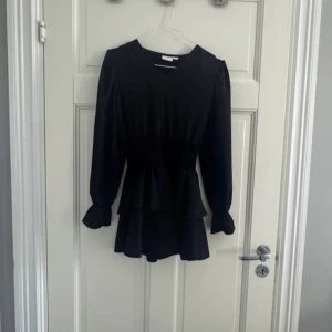 Svart volangklänning Gina Tricot - Supersnygg svart kortklänning från Gina Tricot med långa ärmar, v-ringning och markerad midja. Klänningen har volanger i kjolen och vid ärmsluten, vilket ger en trendig och feminin look. Perfekt för dig som gillar stilrena och coola outfits.