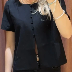 Svart blus med knappar framtill - Stilren svart blus med korta ärmar och raka linjer. Blusen har dekorativa klädda knappar längs hela framsidan och en enkel, klassisk rund halsringning. Perfekt att matcha med jeans eller kjol för en clean look.
