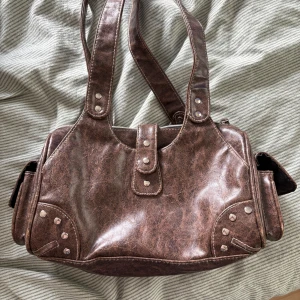 Brun handväska  - Cool brun axelväska i skinnimitation med silvriga nitar och detaljer. Väskan har dubbla axelband, dragkedja upptill och praktiska sidofickor med tryckknapp. Perfekt storlek för vardagens essentials och ger en vintage vibe😽
