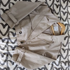 Beige croppad trenchcoat från Bershka - Säljer en Helt ny beige croppad trenchcoat från Bershka med klassisk krage, dubbelknäppning och bälte vid ärmsluten. Jackan har elastisk midja bak och är tillverkad i 100% bomull. Perfekt för dig som vill ha en stilren och trendig look.