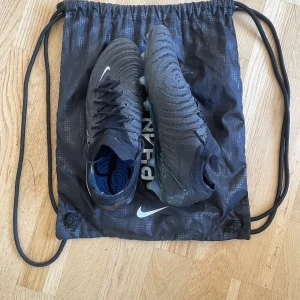 Nike Phantom GT2 Elite fotbollsskor svarta - Säljer ett par svarta Nike Phantom GT2 Elite fotbollsskor med snygg struktur och silvriga detaljer. Skorna har snörning och en tight passform för maximal bollkontroll. Ovandelen är i syntetmaterial och sulan har dobbar för spel på konstgräs.Perfekta för dig som vill dominera på planen.