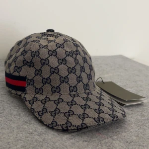 Klassisk gg keps beige - För frågor och video skicka gärna dm. Storlek ”M” Snygg beige keps från Gucci med klassiskt GG-monogram i mörkblått. Kepsen har en röd och marinblå rand på sidan och justerbart spänne bak i svart skinn. Perfekt accessoar för en trendig look.