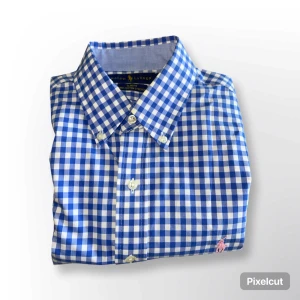 Blårutig skjorta från Ralph Lauren - Klassisk blårutig skjorta från Ralph Lauren med button-down krage och broderad logga på bröstet. Skjortan har långa ärmar och vita knappar framtill. Perfekt för en stilren och tidlös look.
