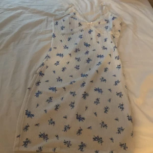 Vit blommig kortklänning med spetsdetaljer - Söt vit kortklänning med blå blommor och tunna axelband. Klänningen har spetskant upptill och är gjord i ett ribbat material. Perfekt för varma dagar när du vill känna dig extra fin. Kan även användas till t.ex skolavslutning eller firande av missommar 