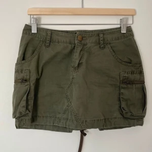 Olivgrön cargo minikjol - Säljer en olivgrön cargo minikjol med fickor och dragkedjor på sidorna. Kjolen har en knapp och dragkedja framtill samt hällor för skärp. 