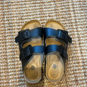 Svarta sandaler med två spännen - Säljer ett par svarta sandaler med två breda remmar och justerbara spännen. Sulan är ergonomiskt formad och har en naturlig beige färg. Perfekta för varma dagar och enkel att matcha med olika outfits.