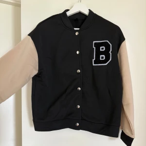 Svart och beige tunn jacka/kofta med B-märke - Säljer en svart tunn jacka/kofta med beige ärmar och ett stort B-märke på bröstet. Jackan har tryckknappar framtill och ribbade muddar vid hals, ärmslut och nederkant. Perfekt för en sportig och trendig look.