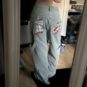 Ljusblå baggy jeans med tryck - Baggy ljusblå jeans med coola tryck på baksidan och ett litet handtryck fram. Byxorna har normal passform i midjan och vida ben. Perfekta för dig som gillar streetstyle och vill sticka ut lite extra.