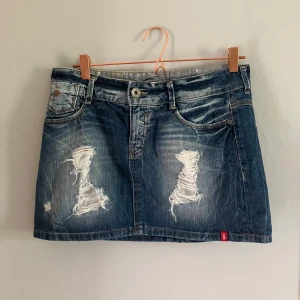 Blå jeanskjol med slitningar - Säljer en blå jeanskjol med slitna detaljer framtill och klassiska bakfickor. Kjolen har en kort passform och stängs med knapp och dragkedja. Pris går att diskutera 